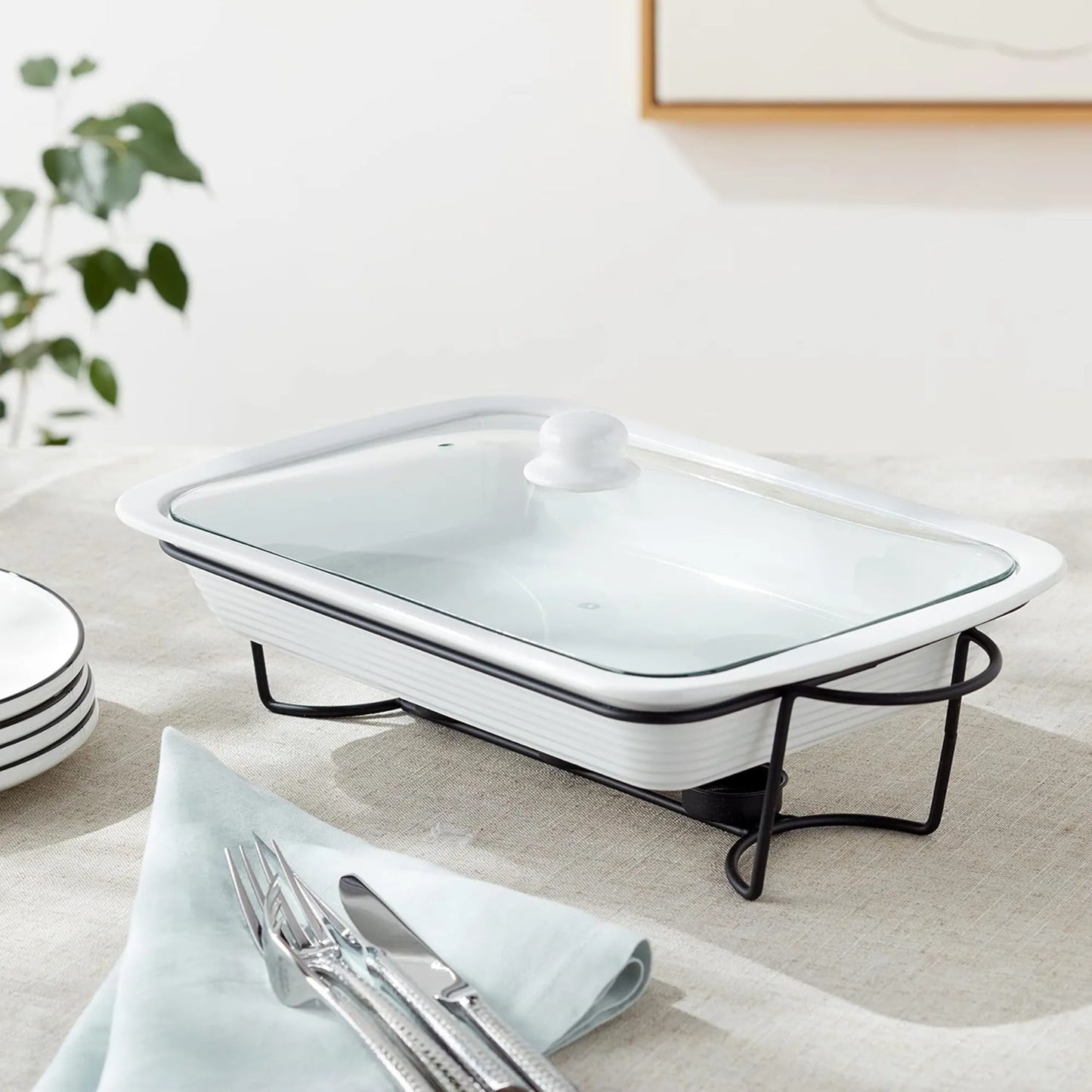 The Le Regalo 3 Piece Bake and Serve Food Warmer Set—a rectangular white ceramic dish with a glass lid on a black metal stand—sits on a table beside napkins, cutlery, and plates, perfect for effortless entertaining. A plant and artwork are in the background.