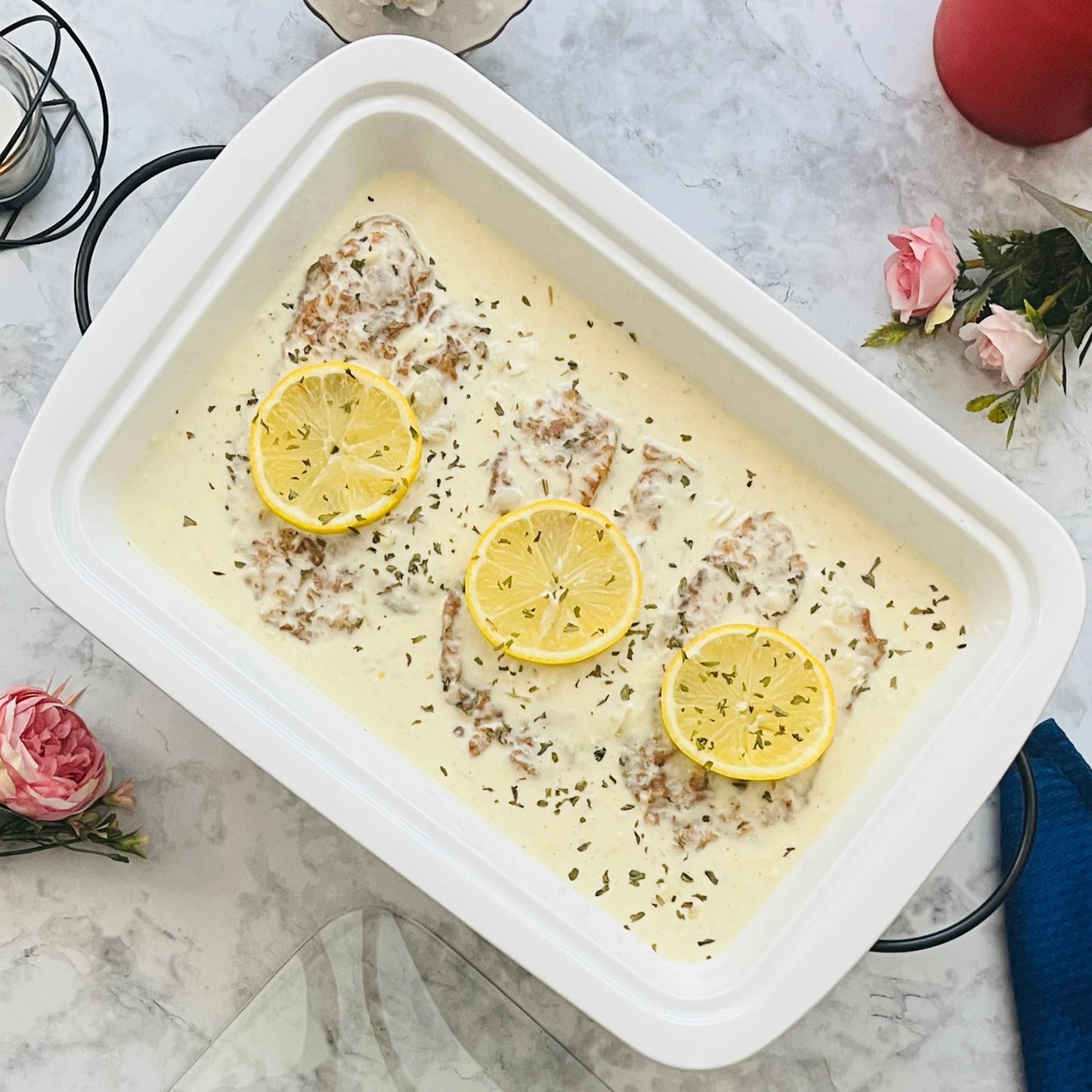 The Le Regalo 3 Piece Bake and Serve Food Warmer Set displays creamy baked chicken breasts topped with lemon and herbs, arranged on a marble surface with pink flowers and utensils for effortless entertaining.
