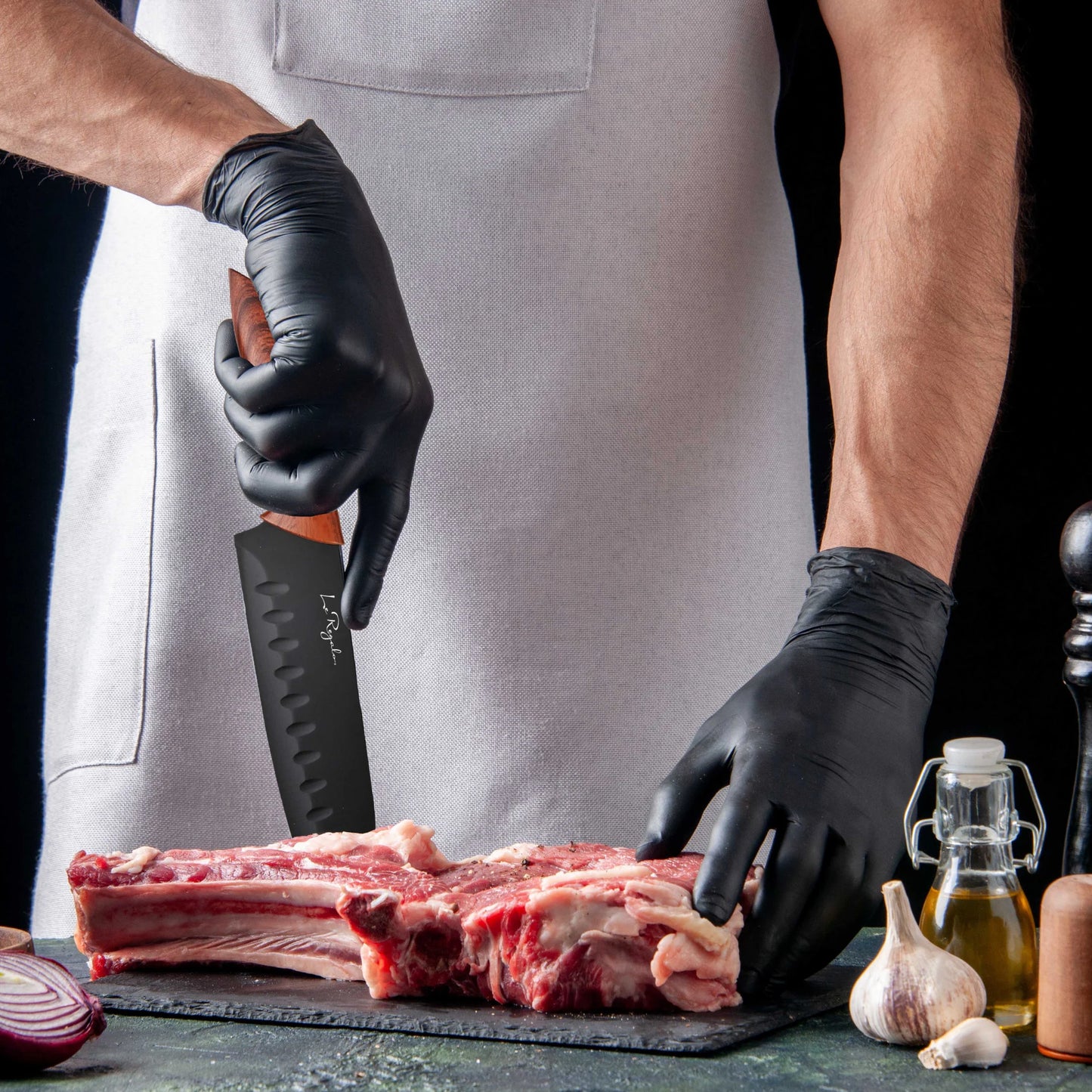 Wearing a white apron and black gloves, a person uses a stainless steel knife to cut raw meat on a black cutting board, with garlic, red onion, oil, a pepper grinder, and the Le Regalo 5 Piece Kitchen Knife Set with Magnetic Holder nearby.