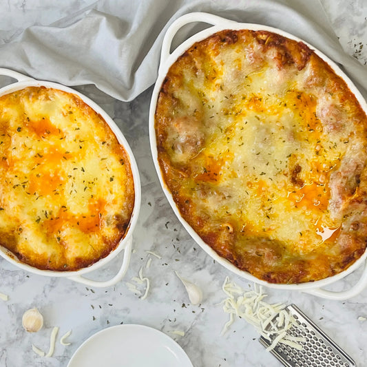 Two golden-brown, cheesy lasagnas bake in the Le Regalo 2 Piece Stoneware Oval Baker Set on marble, topped with herbs. Shredded cheese, garlic cloves, and a grater rest beside the Le Regalo bakers.