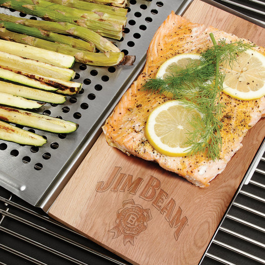 Grilled asparagus, zucchini, and a salmon fillet with lemon and dill cook on the grill using the Jim Beam 2 Piece Grill Topper Set. The cedar plank under the salmon features the Jim Beam brand.