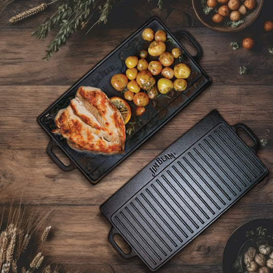 The Jim Beam Cast Iron Reversible Grill/Griddle holds cooked chicken breast and roasted baby potatoes on a wooden table, with wheat and herbs nearby, highlighting the versatility of stovetop cooking with Jim Beam.