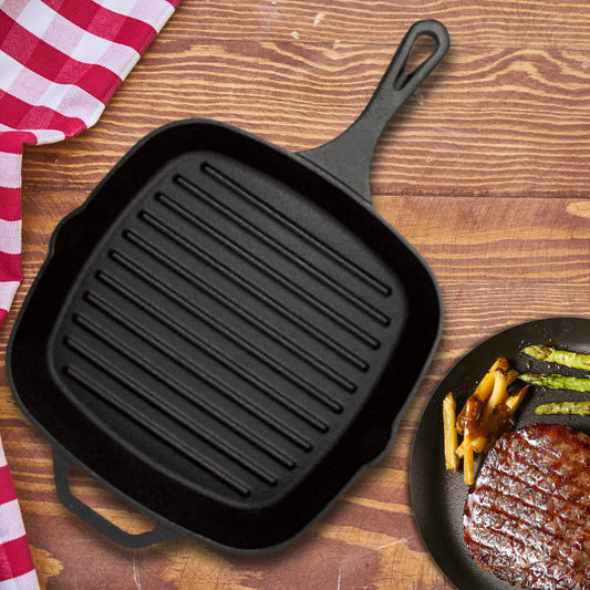 The Jim Beam Cast Iron Square Grill Pan, known for excellent heat retention, rests on a wooden surface beside a red and white checkered cloth and a plate of steak, asparagus, and roasted potatoes.