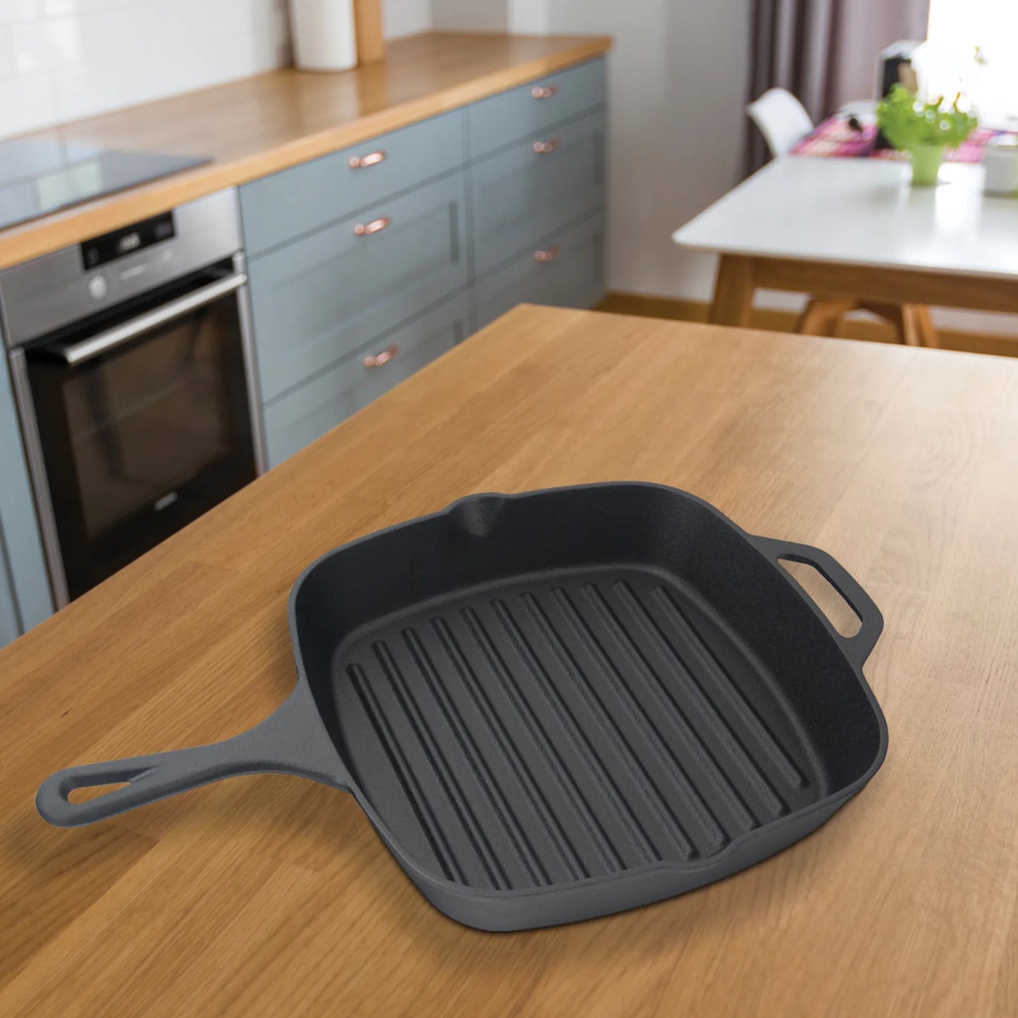 The Jim Beam Cast Iron Square Grill Pan, known for excellent heat retention, sits on a wooden kitchen counter in a modern kitchen with blue cabinets and a dining table—ideal for grilling enthusiasts.