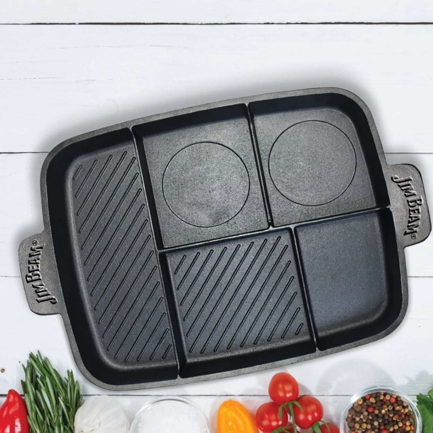 The Jim Beam Cast Iron 5-in-1 Skillet sits on a white surface, surrounded by cherry tomatoes, bell peppers, rosemary, and small bowls of salt and peppercorns. Its versatile design features both grill and griddle surfaces with ridged and flat sections.