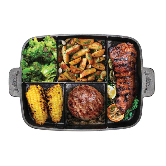 A Jim Beam Cast Iron 5-in-1 Skillet holds grilled broccoli, roasted potatoes, corn, a hamburger patty, and a seasoned steak with lemon and herbs, all arranged in divided sections and viewed from above on a white background.