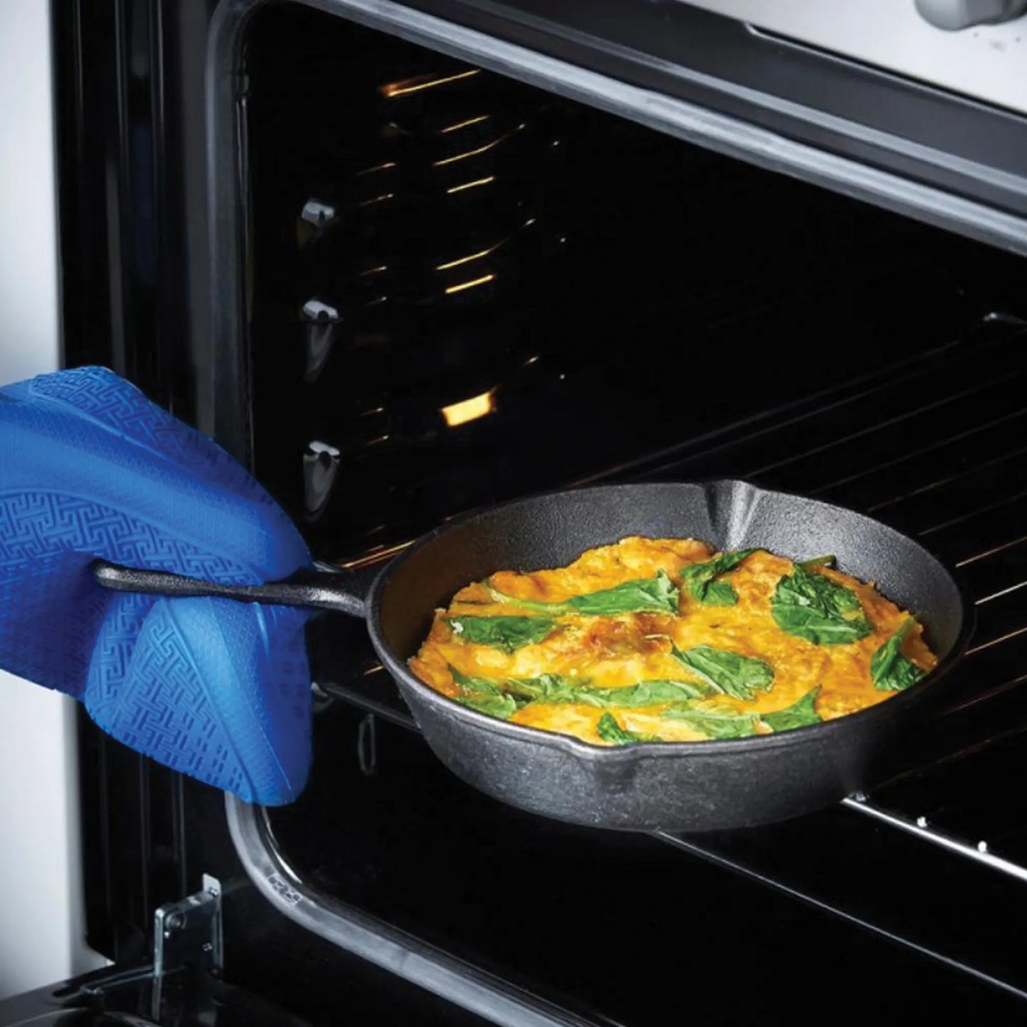 Using a blue oven mitt, someone is placing or removing a baked egg and spinach dish in the Jim Beam Cast Iron 3 Piece Skillet Set, highlighting the versatility of pre-seasoned cookware from Jim Beam.