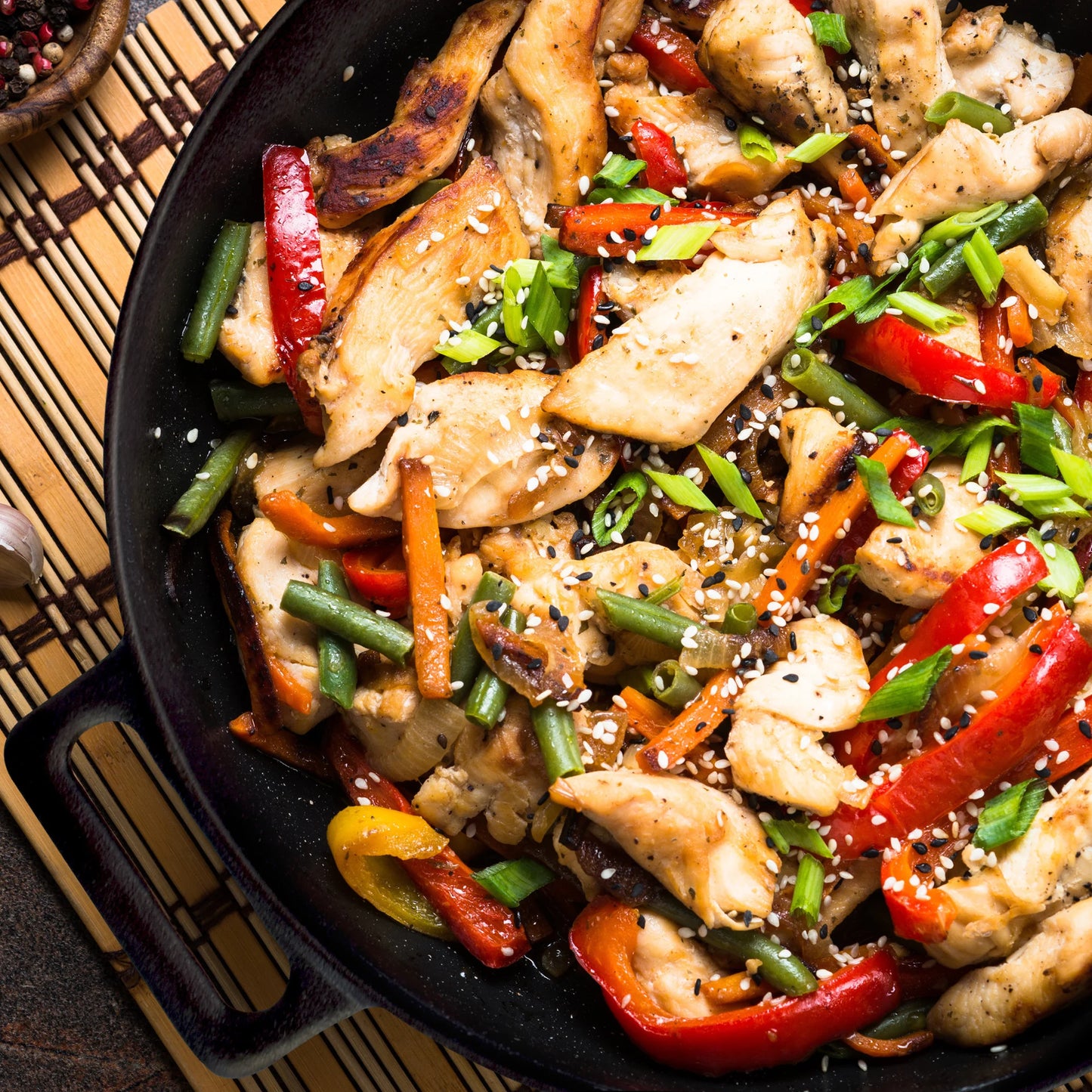 A vibrant chicken stir-fry with red bell peppers, green beans, carrots, onions, and sesame seeds, garnished with sliced green onions and served in the Jim Beam 12 Cast Iron Wok by Jim Beam on a woven placemat.
