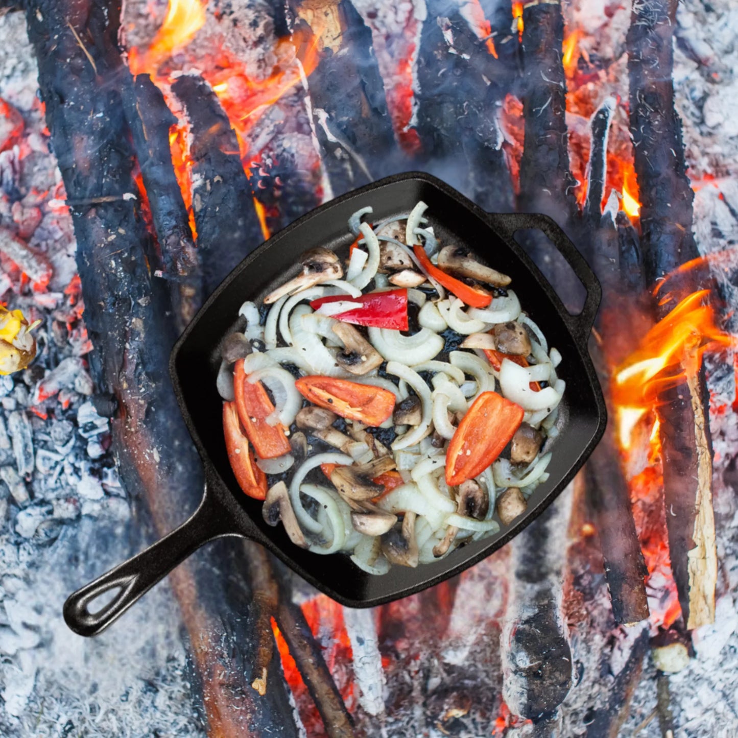 The Jim Beam Cast Iron 10.5 Skillet is shown over an open campfire, filled with sliced onions, mushrooms, and red bell peppers as flames and burning logs cook the vegetables to perfection.