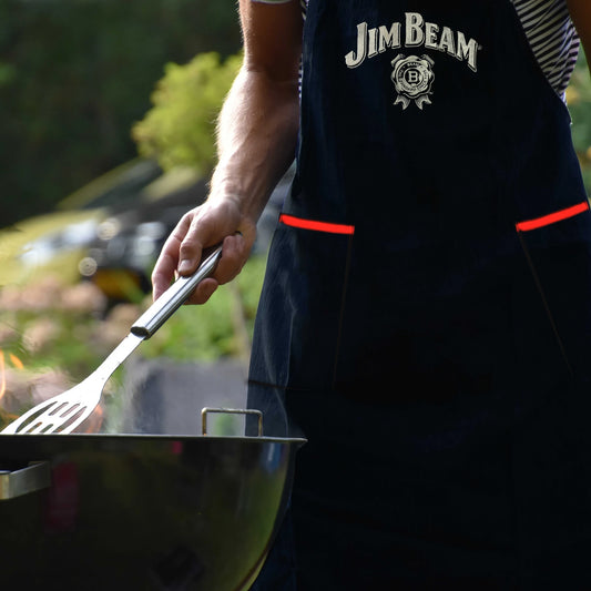 Someone grills outdoors wearing the Jim Beam Classic Grilling Apron, holding a spatula at the barbecue, with greenery in the background.
