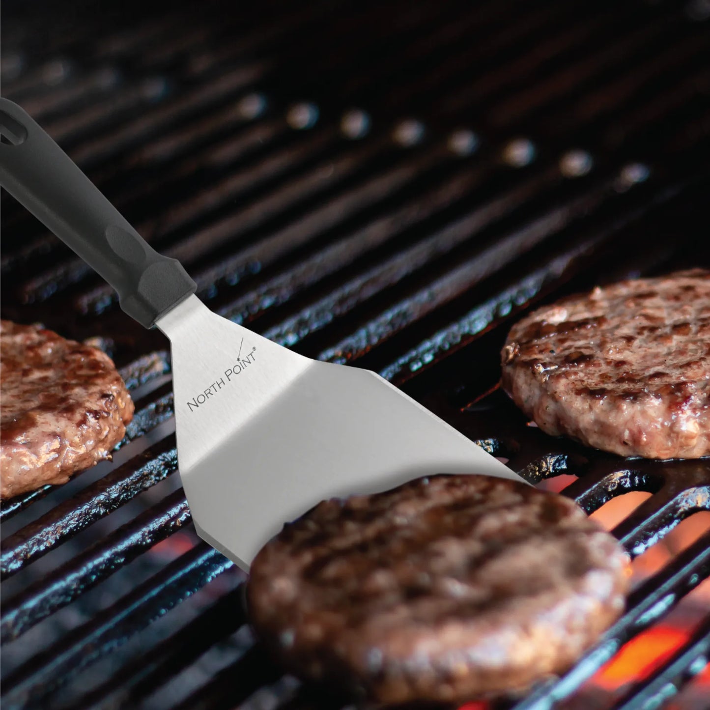 A metal spatula from the North Point 3 Piece Burger Kit flips a beef patty on the grill alongside three others, each perfectly shaped with the North Point press, showing grill marks above glowing coals.