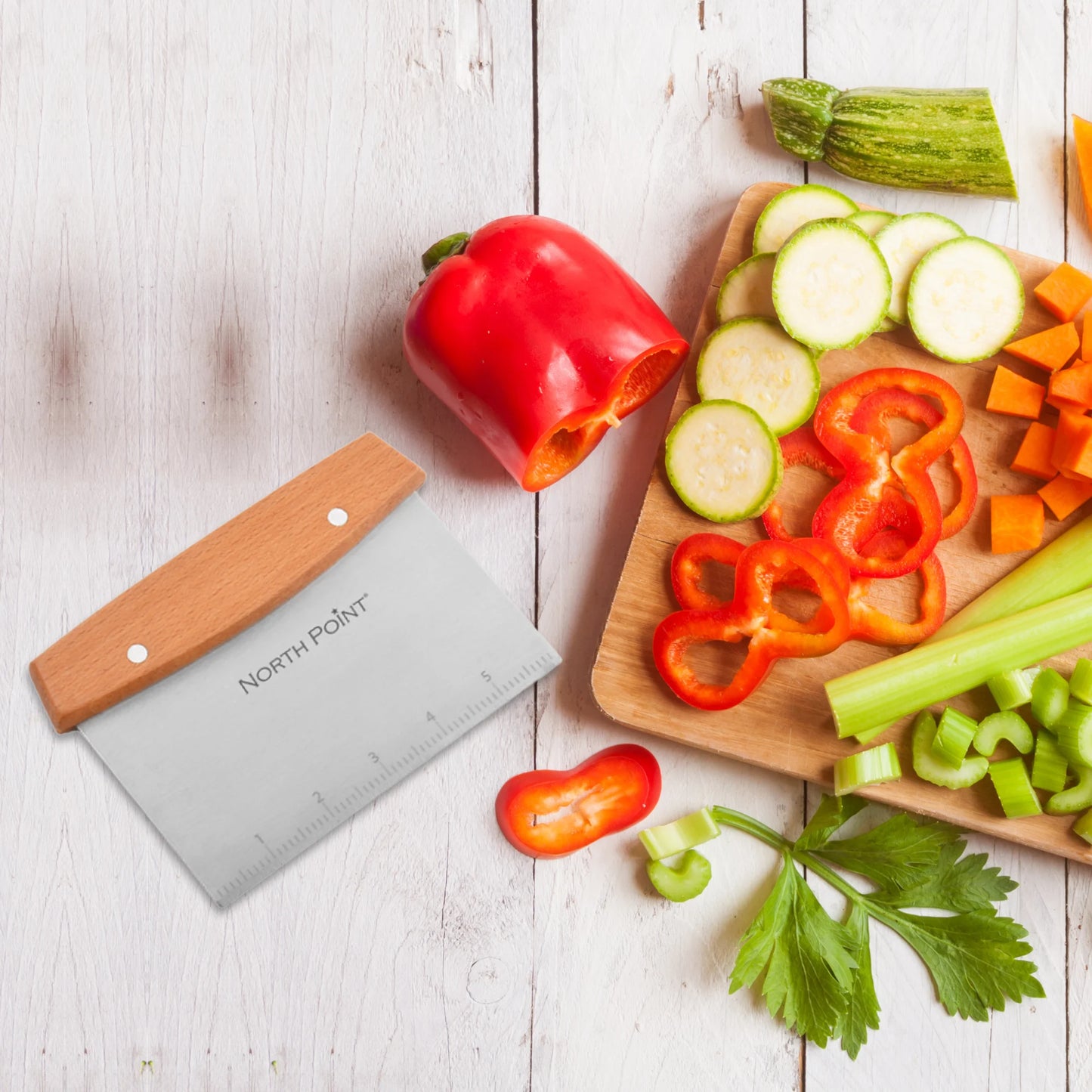 A wooden cutting board with sliced zucchini, red bell pepper, carrots, and celery sits on a white wooden surface. A North Point dough scraper from the North Point 8 Piece Griddle Set rests beside the board.