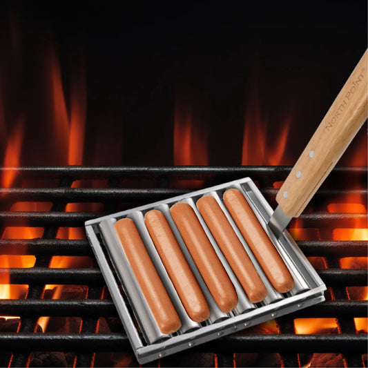 Five hot dogs are being grilled over an open flame on the North Point Hot Dog Roller which has stainless steel construction, a wooden handle.