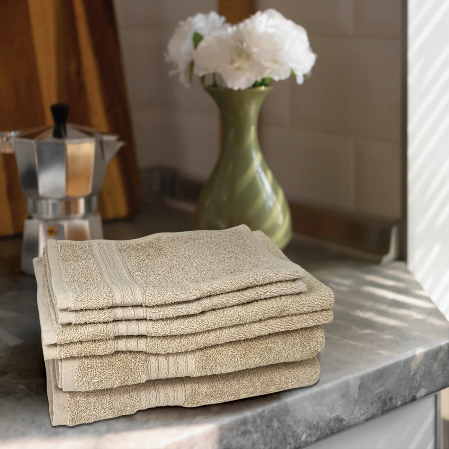 A stack of neatly folded Textilia Set of 6 Bath Towel Set – Beige sits on a gray kitchen countertop, with a green vase of white flowers and a silver espresso maker softly lit by warm sunlight in the background.