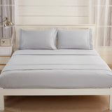 A neatly made bed features the Textilia Bedsheet Set by Textilia in light gray brushed microfiber, styled with two pillows, bright white walls, large windows, a lamp, and a small nightstand for luxurious comfort.