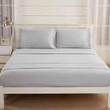 A neatly made bed features the Textilia Bedsheet Set in light gray brushed microfiber with two pillows, set against a white wooden headboard. A nightstand with a lamp and decor sits on the left, and large windows have sheer white curtains.