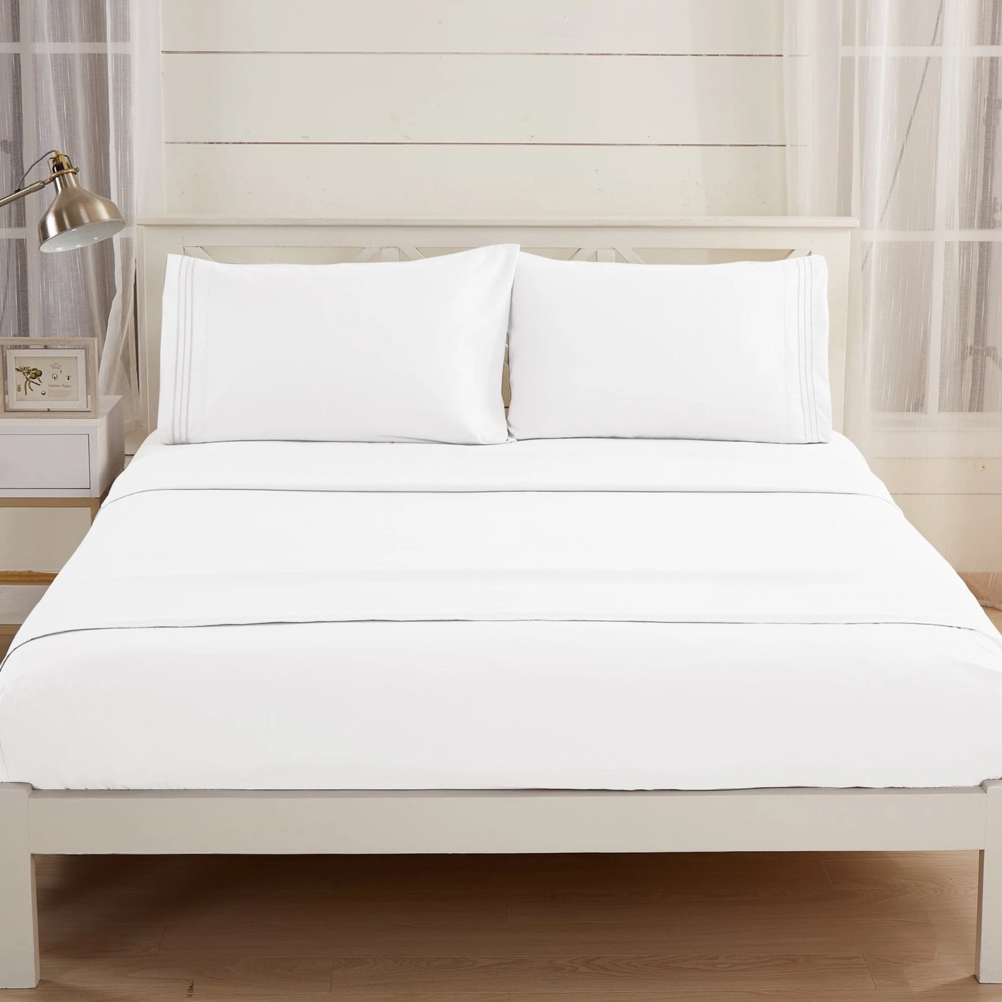 A neatly made bed with Textilia Bedsheet Set by Textilia and white pillows sits in a bright room with white walls, large windows, a nightstand, lamp, and frame.