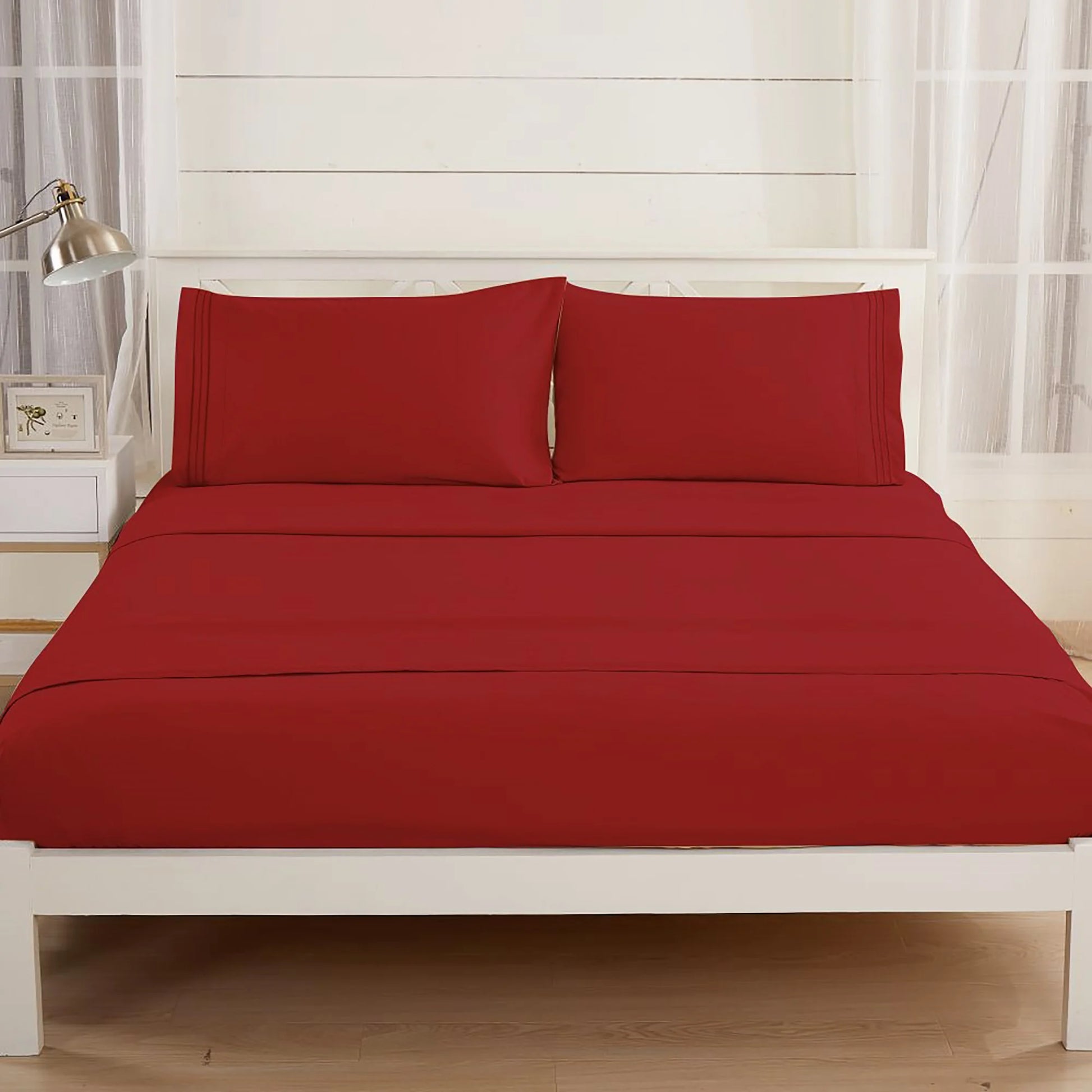 A neatly made bed features the Textilia Bedsheet Set by Textilia in burgundy brushed microfiber, paired with matching pillowcases on a white frame. The bright room includes light walls, large windows with sheer curtains, a lamp, and a frame.