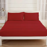 A neatly made bed features the Textilia Bedsheet Set by Textilia in burgundy brushed microfiber, paired with matching pillowcases on a white frame. The bright room includes light walls, large windows with sheer curtains, a lamp, and a frame.