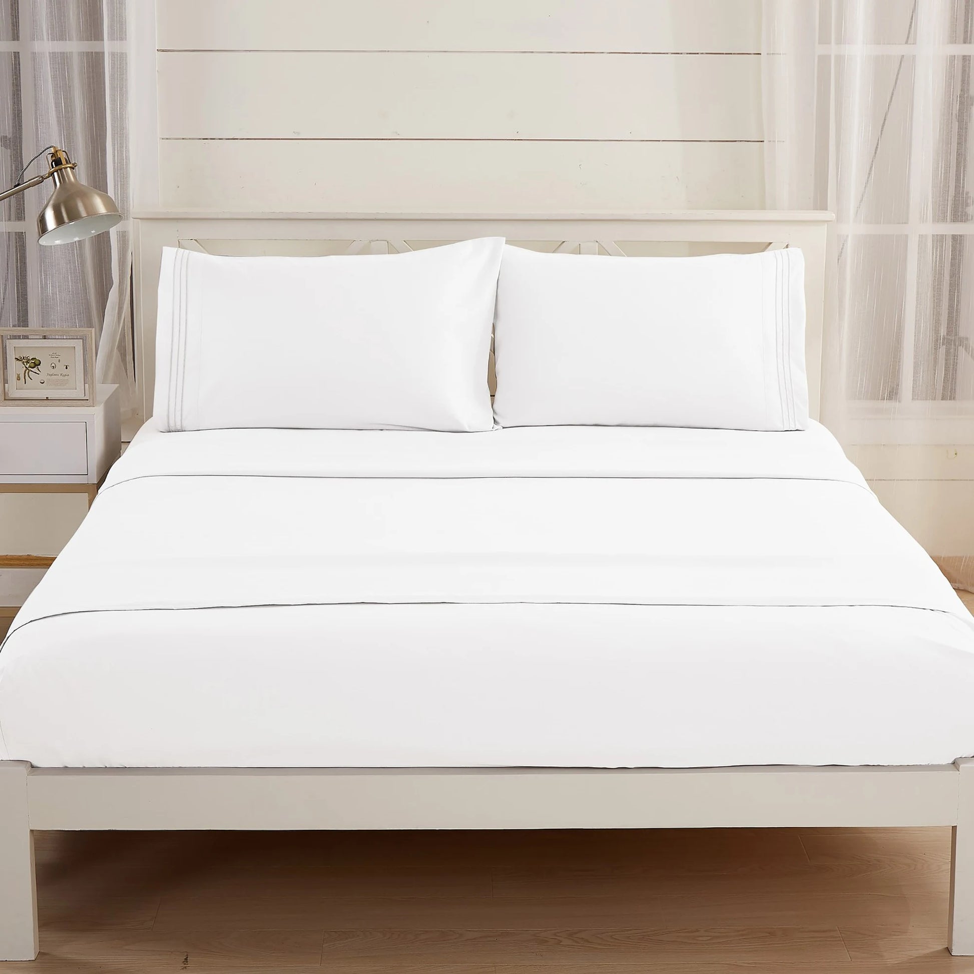 A neatly made bed features crisp white Textilia Bedsheet Set by Textilia and two white pillows, set against a light wall with large windows and sheer curtains. A small nightstand with a lamp and frame sits beside the bed.