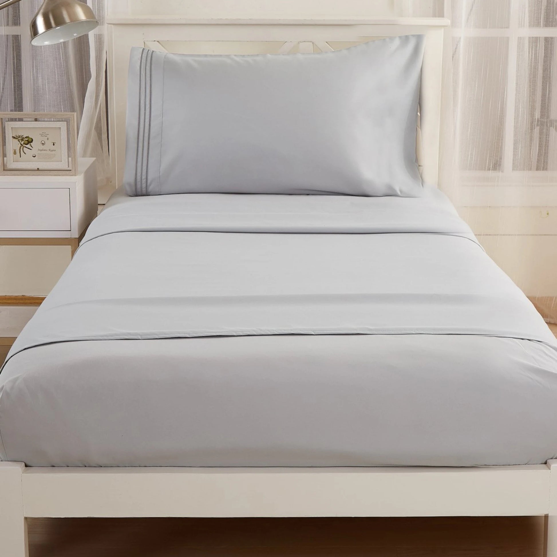 A single bed is dressed in the Textilia Bedsheet Set by Textilia—light gray brushed microfiber with a matching pillowcase and sheet. Set against a white headboard, its beside a nightstand with a lamp and a frame.