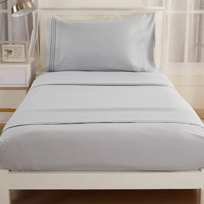 A single bed is dressed in the Textilia Bedsheet Set by Textilia—light gray brushed microfiber with a matching pillowcase and sheet. Set against a white headboard, its beside a nightstand with a lamp and a frame.