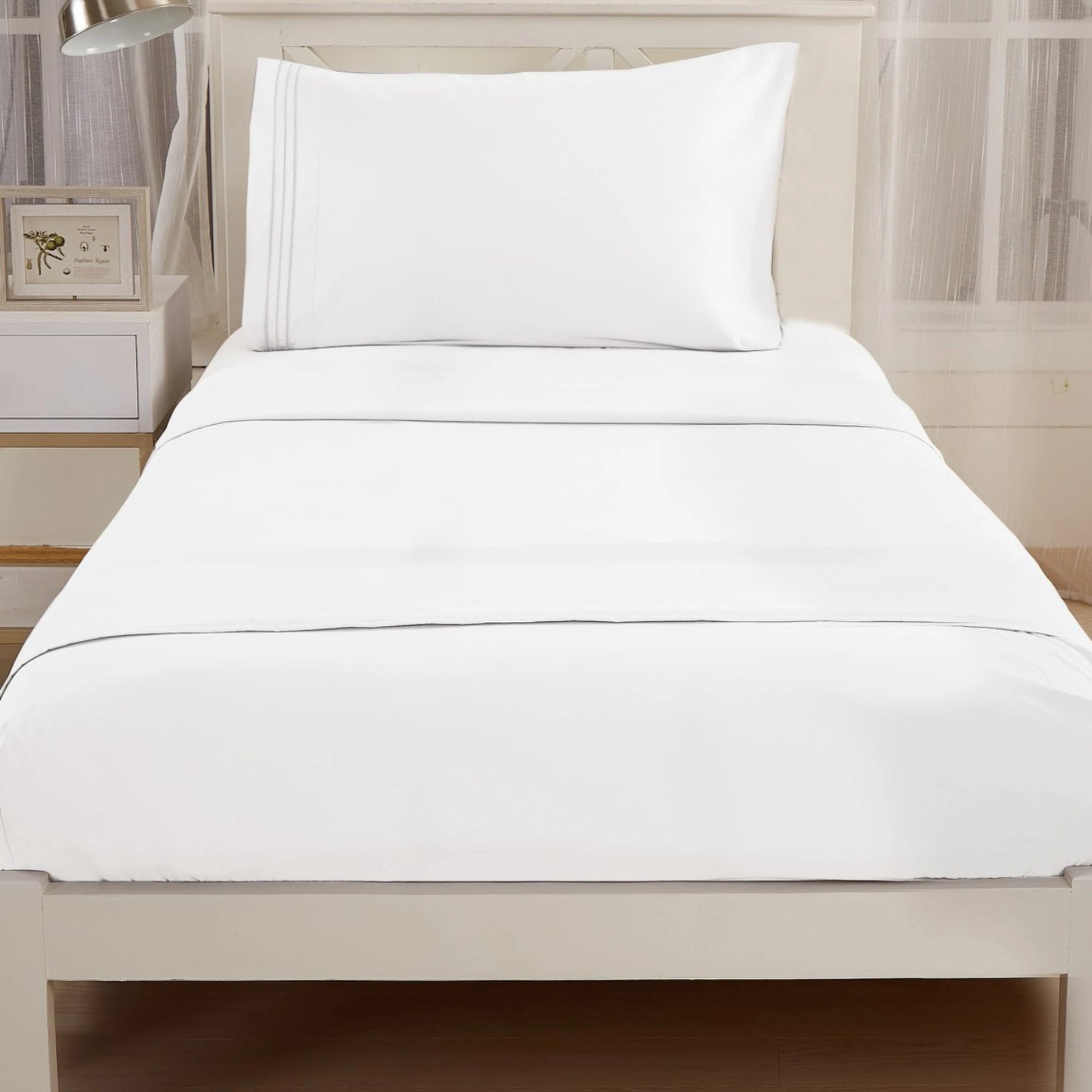 A single bed with the Textilia Bedsheet Set by Textilia, featuring white hypoallergenic sheets and a pillow, is in a bright room with wooden floors, a nightstand, and a framed picture to the left.