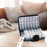 The Smart Essentials 14-Day Pill & Vitamin Organizer in black, featuring labeled morning, evening, and bedtime compartments, is open on a wooden table in a cozy living room with a beige sofa and blankets in the background.