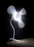 The North Point Clip Light with Fan 11W, a white flexible gooseneck desk fan lamp by North Point, is clipped to the edge of a wooden surface with spinning blades and an integrated LED against a dark background.