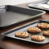 Two Le Regalo baking trays from the Set of 3 rest on a wooden surface—one with six chocolate chip cookies, the other empty. In the background, white cloths are stacked neatly.