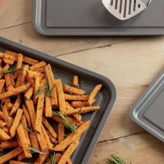 A Le Regalo Set of 3 Baking Tray, filled with seasoned sweet potato fries and fresh rosemary, rests on a wooden surface alongside two other Le Regalo trays and a metal spatula.