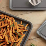 A Le Regalo Set of 3 Baking Tray, filled with seasoned sweet potato fries and fresh rosemary, rests on a wooden surface alongside two other Le Regalo trays and a metal spatula.