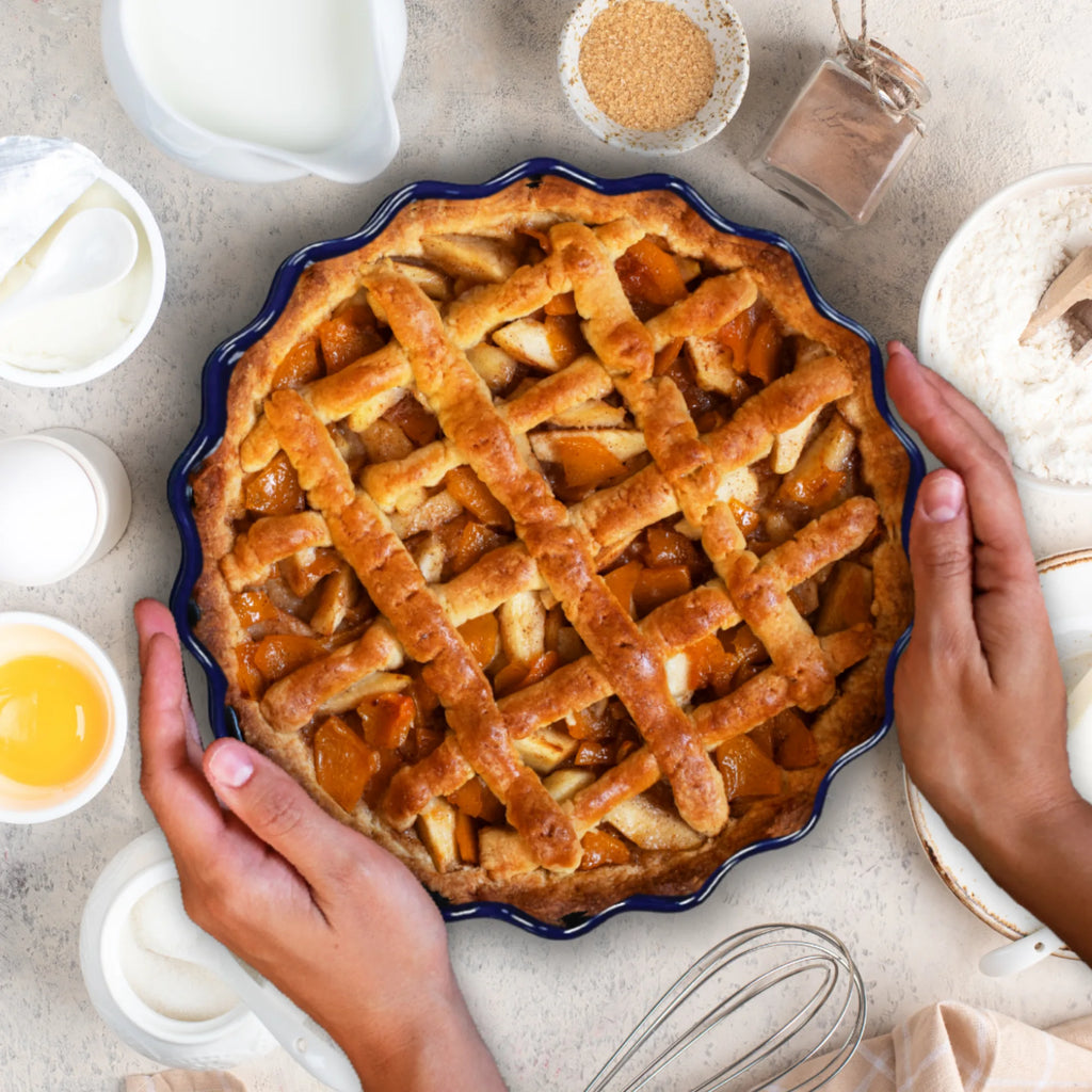 A person holds a freshly baked lattice-topped apple pie in a Le Regalo Stoneware Pie Dish, surrounded by eggs, sugar, flour, milk, and kitchen utensils on a light-colored surface.