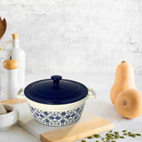 The Le Regalo Stoneware Casserole with Lid, featuring a blue top and floral design, rests on a wooden cutting board beside a butternut squash, pumpkin seeds, a pepper grinder, white bottles, and a mortar and pestle.