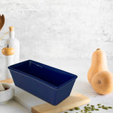 The Le Regalo Stoneware Loaf Pan in blue rests on a wooden board, surrounded by butternut squash, pumpkin seeds, white bottles, utensils, and a pepper grinder—highlighting Le Regalo’s kitchen versatility on a bright countertop.