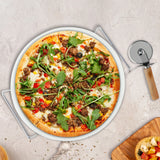 A freshly baked pizza rests on the Le Regalo 3 Piece Pizza Stone Set by Le Regalo, shown with a pizza cutter and wooden board holding a small bowl of food, all displayed on a light-colored surface.