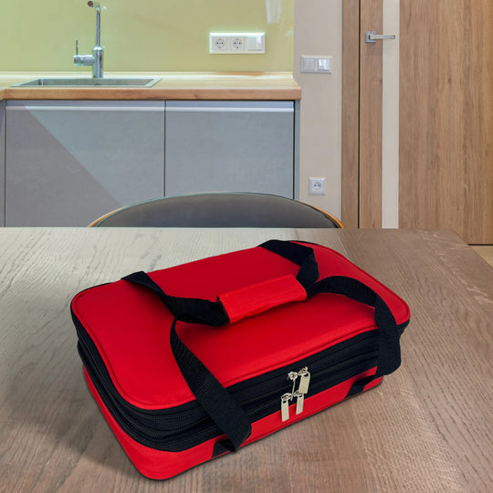 The Le Regalo 5 Piece Bake & Take Set, a red soft-sided insulated carrier with black handles, is placed on a wooden table in a modern kitchen featuring a sink and light wood cabinets.