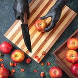 Wearing black gloves, a person slices a red apple with the Le Regalo 5 Piece Kitchen Knife Set with Magnetic Holder on a striped wooden cutting board. Whole apples and flower petals rest on a green countertop and in a wooden box.