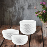 The Le Regalo 4 Piece Stoneware Bowl Set, featuring a subtle honeycomb pattern in white, is stacked on a rustic wooden surface beside a basket of wildflowers, set against a textured gray wall.