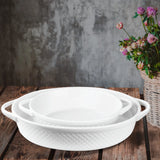 The Le Regalo 2 Piece Stoneware Oval Baker Set, featuring two white handled bakers nested together, sits on a rustic wooden table beside a wicker basket of colorful flowers. A textured gray wall completes the background.