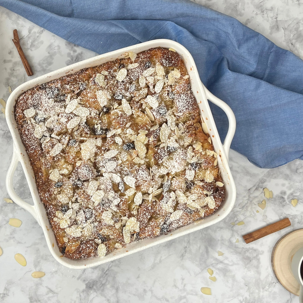 The Le Regalo 10 Stoneware Square Baker, in white, holds a golden-brown dessert topped with flaked almonds and powdered sugar. It sits on a marble surface beside a blue cloth and cinnamon sticks.
