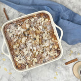 The Le Regalo 10 Stoneware Square Baker, in white, holds a golden-brown dessert topped with flaked almonds and powdered sugar. It sits on a marble surface beside a blue cloth and cinnamon sticks.