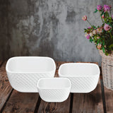 The Le Regalo 3 Piece Stoneware Square Bowl Set is displayed in three sizes on a rustic wood surface, with a wicker basket of colorful flowers to the right, showcasing this versatile Le Regalo kitchen accessory.