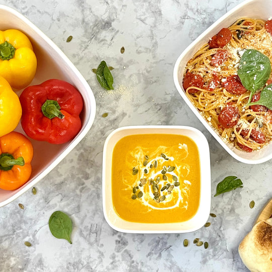 A bowl of creamy orange soup in the Le Regalo 3 Piece Stoneware Square Bowl Set, topped with seeds and cream, sits on marble surrounded by bell peppers, spaghetti with tomatoes and basil, spinach leaves, and bread—a stylish Le Regalo accessory.