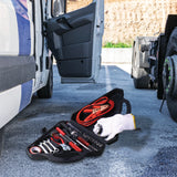 An open American Builder 33 Piece Roadside Emergency Kit with tools, jumper cables, and white gloves is on the ground between a white van and a truck parked on asphalt, with a dirt embankment in the background.