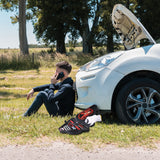 A person sits on grass by a white car with the hood open, talking on the phone. An American Builder 33 Piece Roadside Emergency Kit by American Builder is nearby, suggesting a car breakdown or roadside emergency.