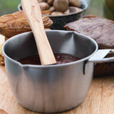The Jim Beam Stainless Steel Basting Pot Set, featuring a wooden spoon and filled with brown sauce, sits on a wooden surface with baked potatoes and a bowl of mushrooms in the background.