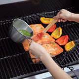 Using the Jim Beam Stainless Steel Basting Pot Set, someone brushes marinade onto salmon fillets in a grill basket, while red and yellow bell pepper halves cook alongside on the grill.