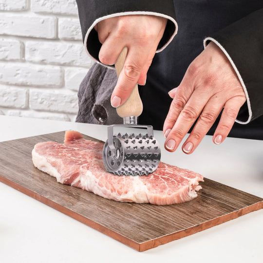 A person uses the Jim Beam Stainless Steel Rolling Meat Tenderizer by Jim Beam to tenderize raw meat on a wooden cutting board, with a white brick wall in the background.