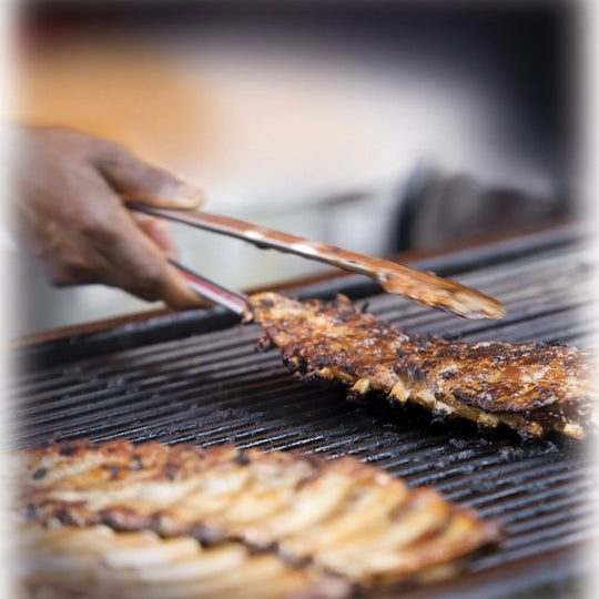 A close-up of a hand using tongs from the Jim Beam 3 Piece Grill Tool Set to grill seasoned ribs on an outdoor barbecue, with smoke rising and a blurred background.